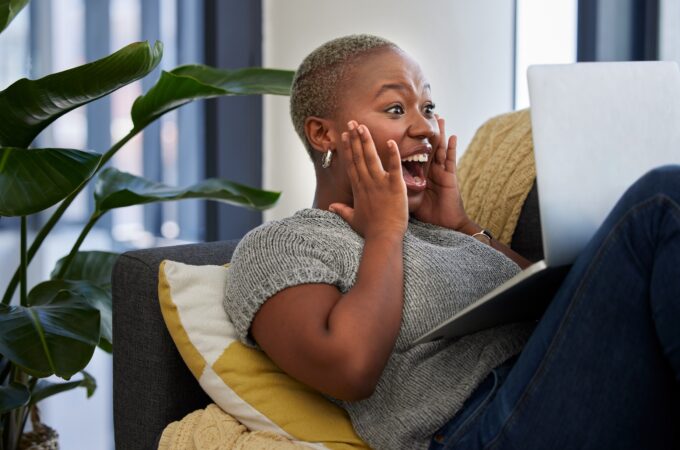 Laptop, surprise and excited woman on sofa shocked with win of digital lottery, competition or award. Wow news, prize winner and girl on gambling website play online poker, bingo or casino game.
