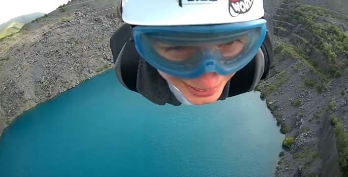 This image shows Zoe during her zipline challenge. Zoe's head is visible and she is wearing a helmet and goggles. Below her is an area of water surrounded by rocks. The location is Penrhyn Quarry in North Wales.