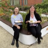 2 staff enjoying some BBQ food together sitting on a wall.