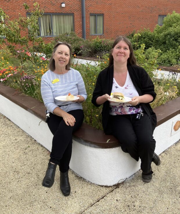 2 staff enjoying some BBQ food together sitting on a wall.