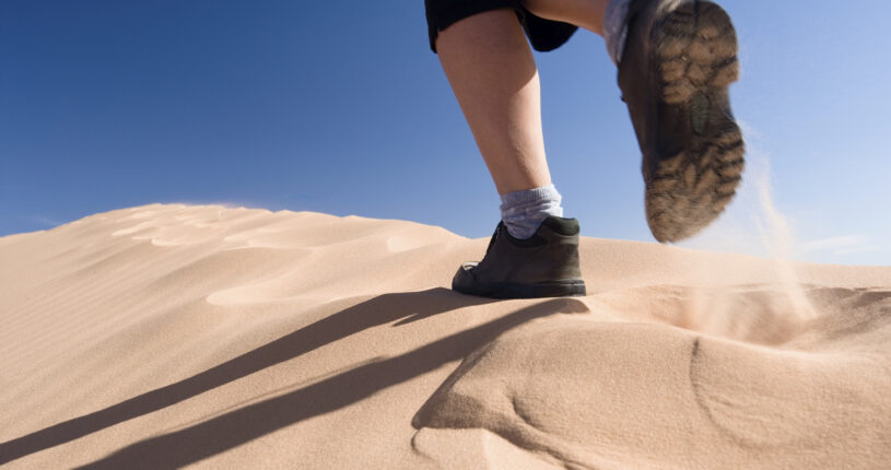 Close-up of a trekker's boots climbing a sand dune, with sand blowing from each step in the Sahara Desert.